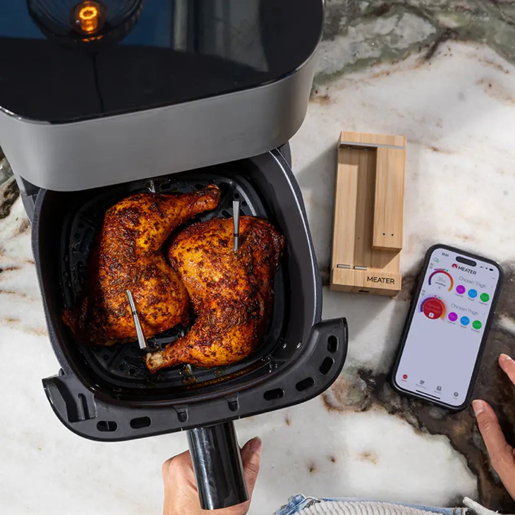 Air fryer with roasted chicken inside, next to a smartphone displaying an app on a marble countertop.