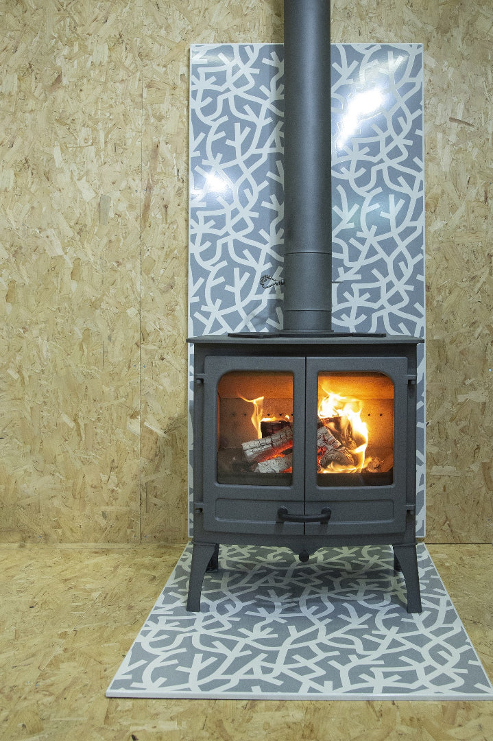 A freestanding Charnwood Island 2 fireplace with a black body and a visible flame, placed on a hearth pad with a geometric pattern. The surrounding area is wooden, and there is a Schott Robax glass panel on the fireplace.