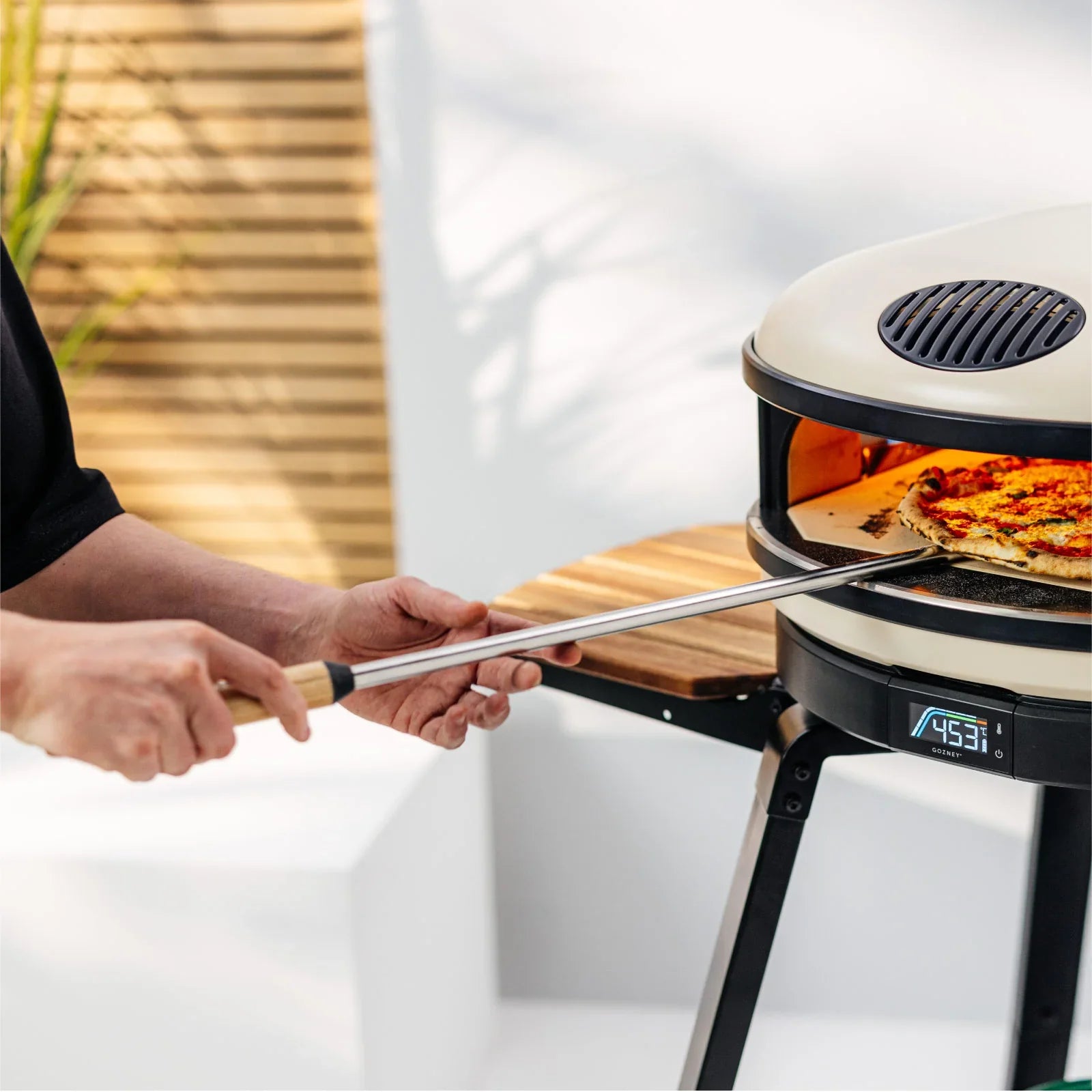Person using a pizza peel to take a pizza out of an outdoor pizza oven.