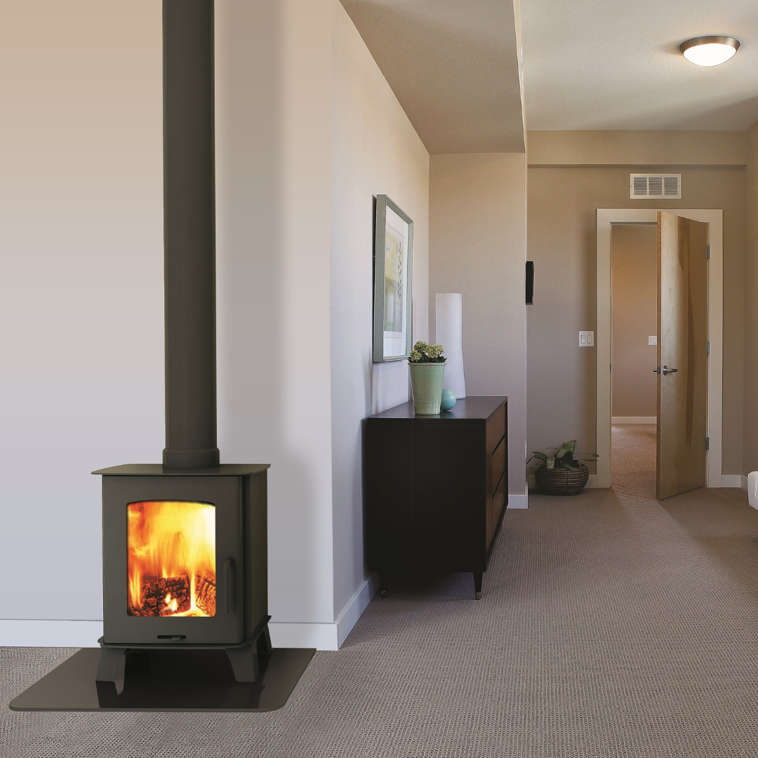 A freestanding wood stove with visible flames in a room with a high ceiling and a door in the background.