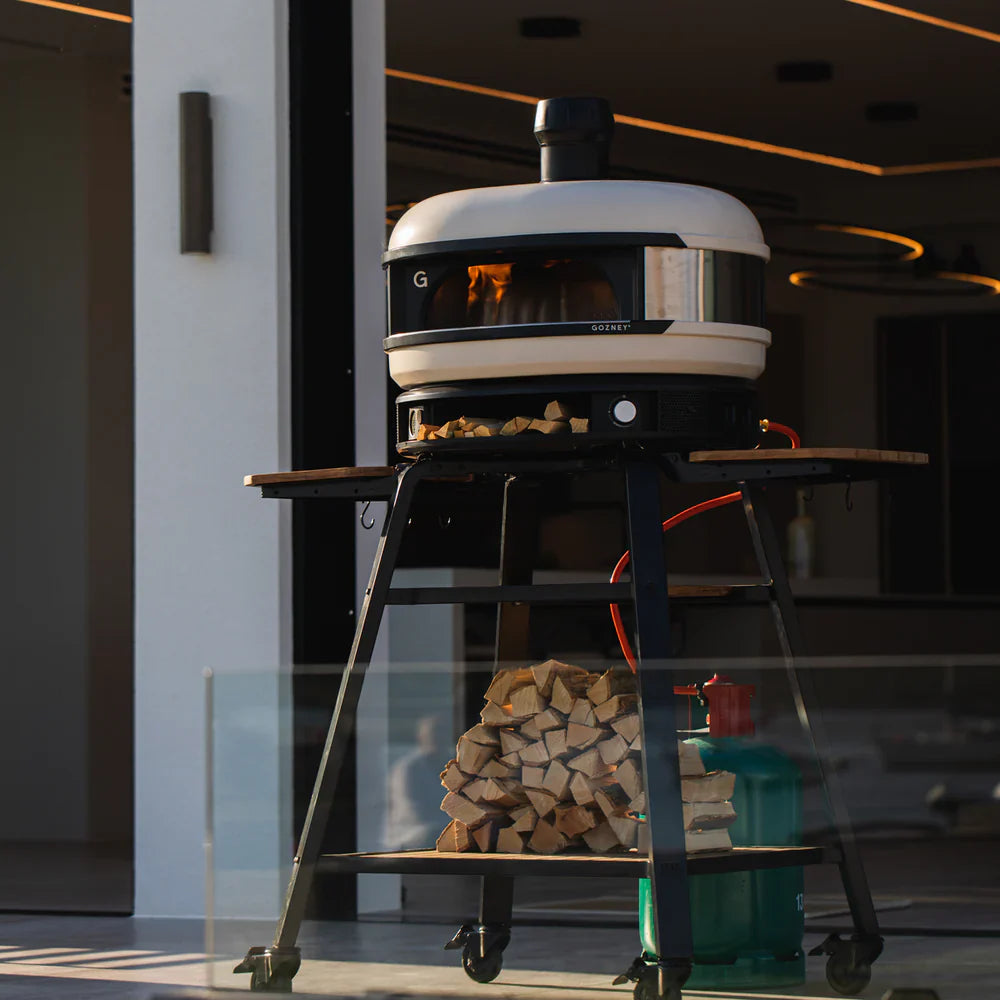 Portable wood-fired pizza oven on a stand with stacked firewood in an indoor setting.