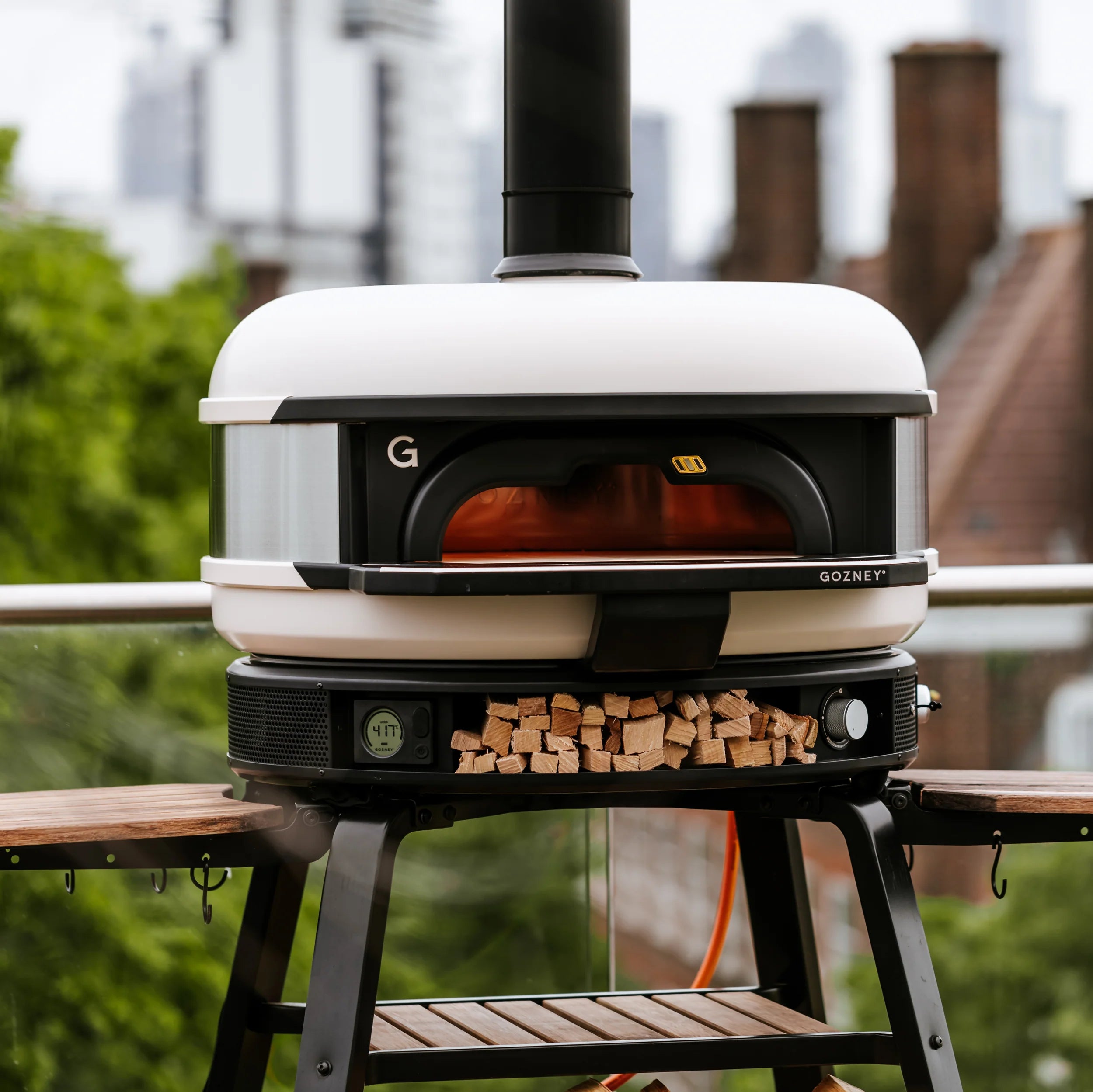 Portable outdoor pizza oven with wood storage on a stand, set against an urban background.