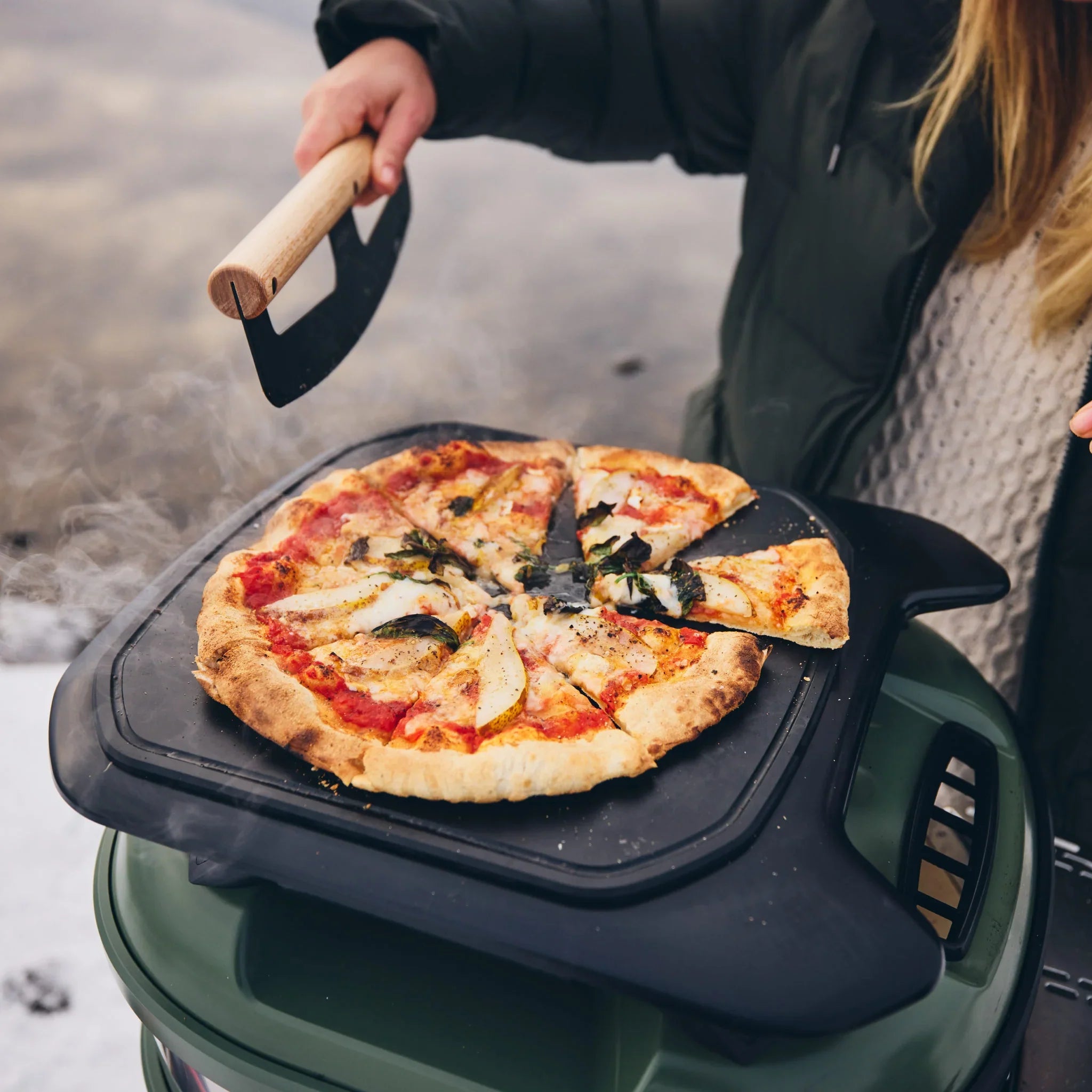 Person holding a pizza on a portable pizza oven in an outdoor setting
