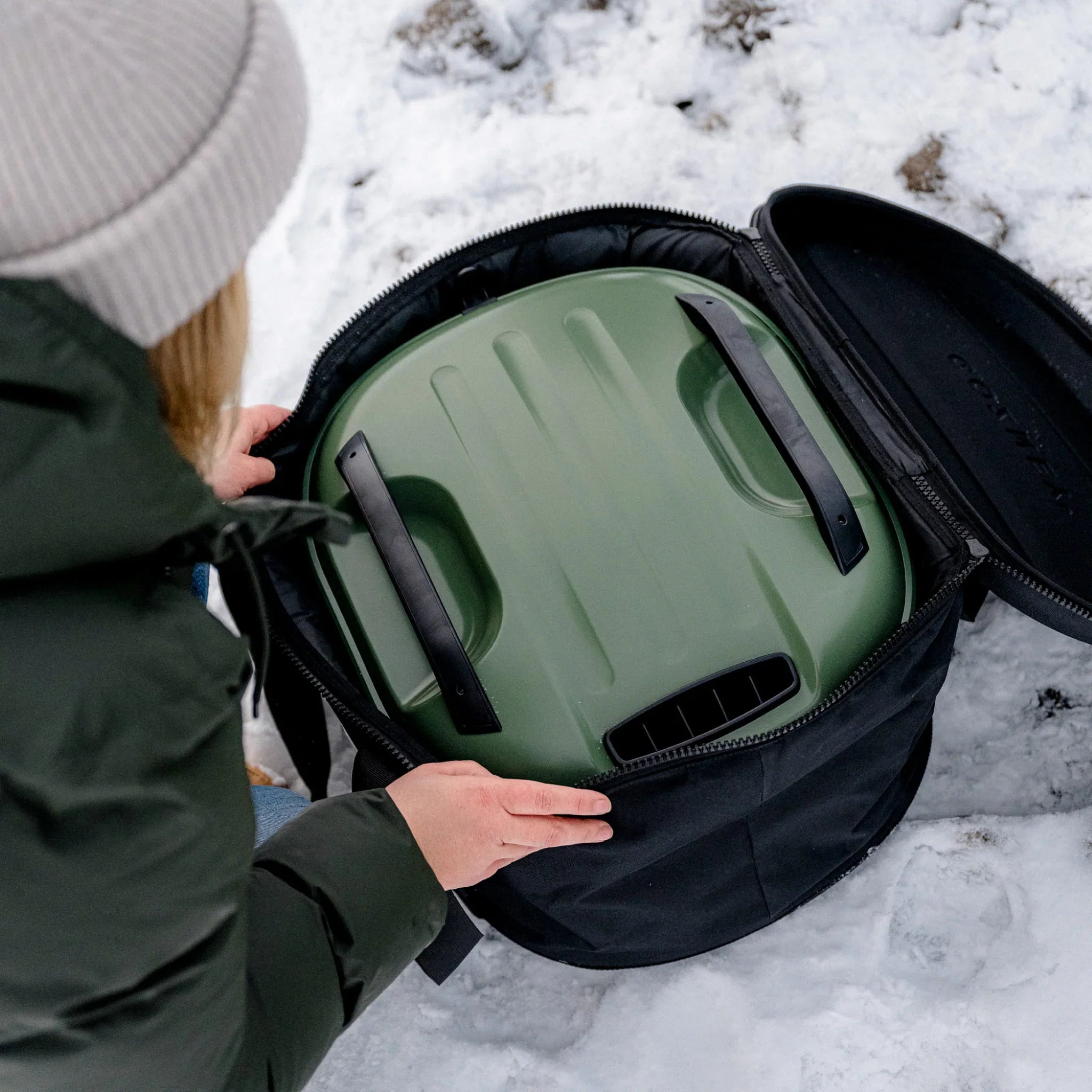 Person opening a black backpack with a green helmet inside on a snowy ground