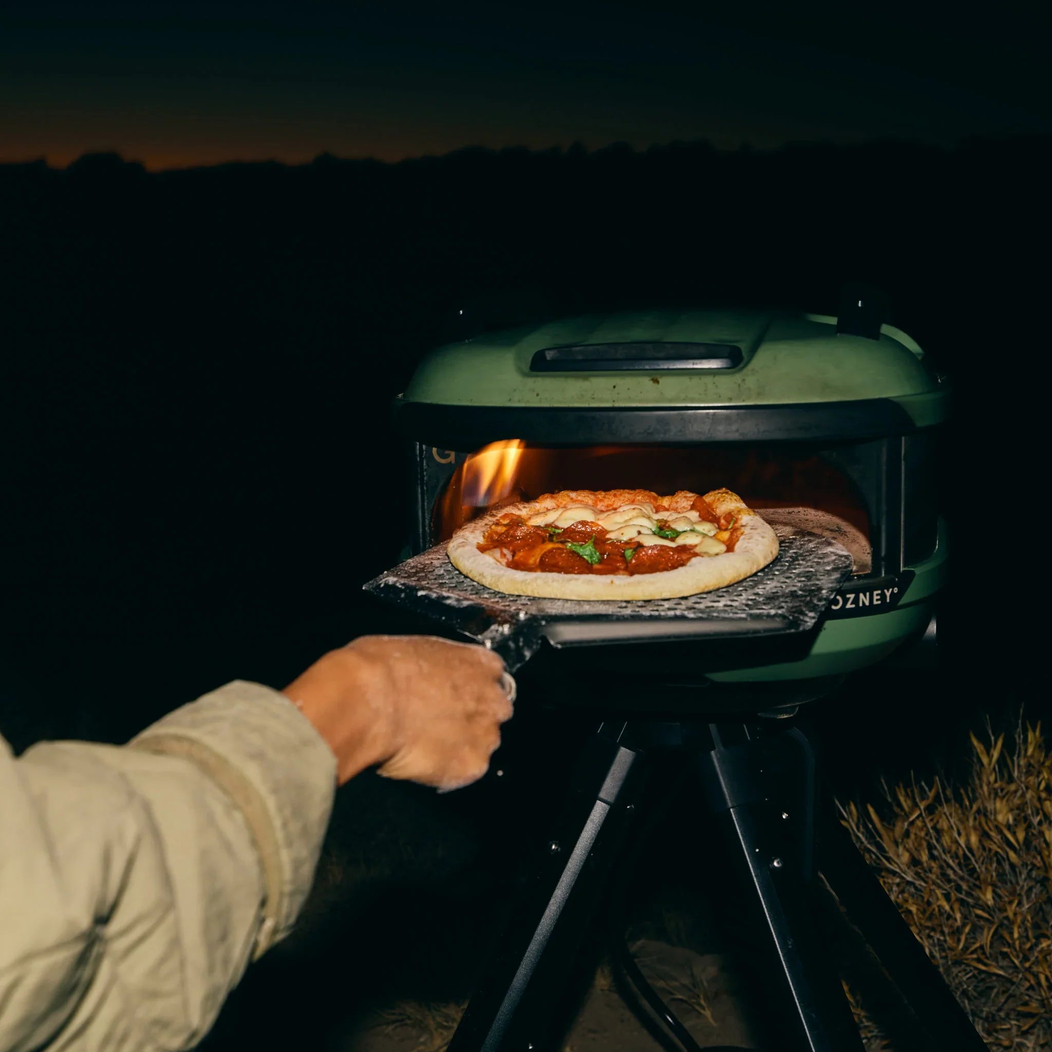 Person cooking a pizza on a portable outdoor oven at night.