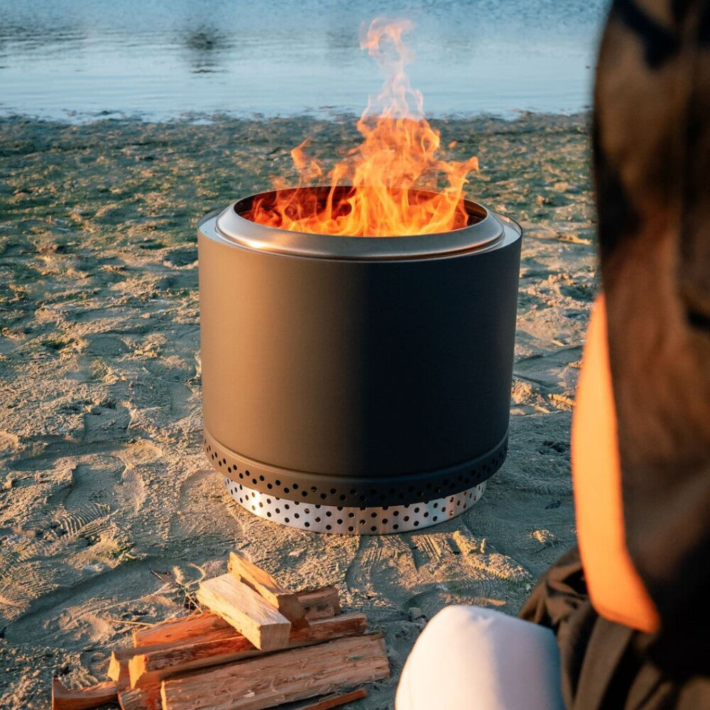 Person sitting by a portable fire pit on a sandy beach with flames burning.