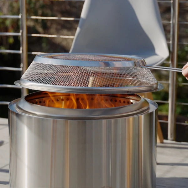 Stainless steel fire pit with a mesh cover on flames, outdoors.