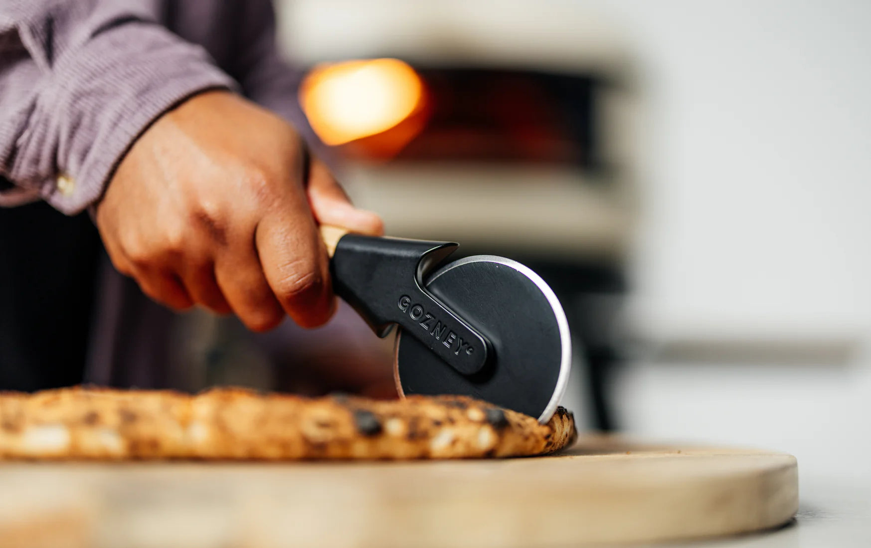 Person using a pizza cutter to slice a pizza on a wooden board with a blurred background