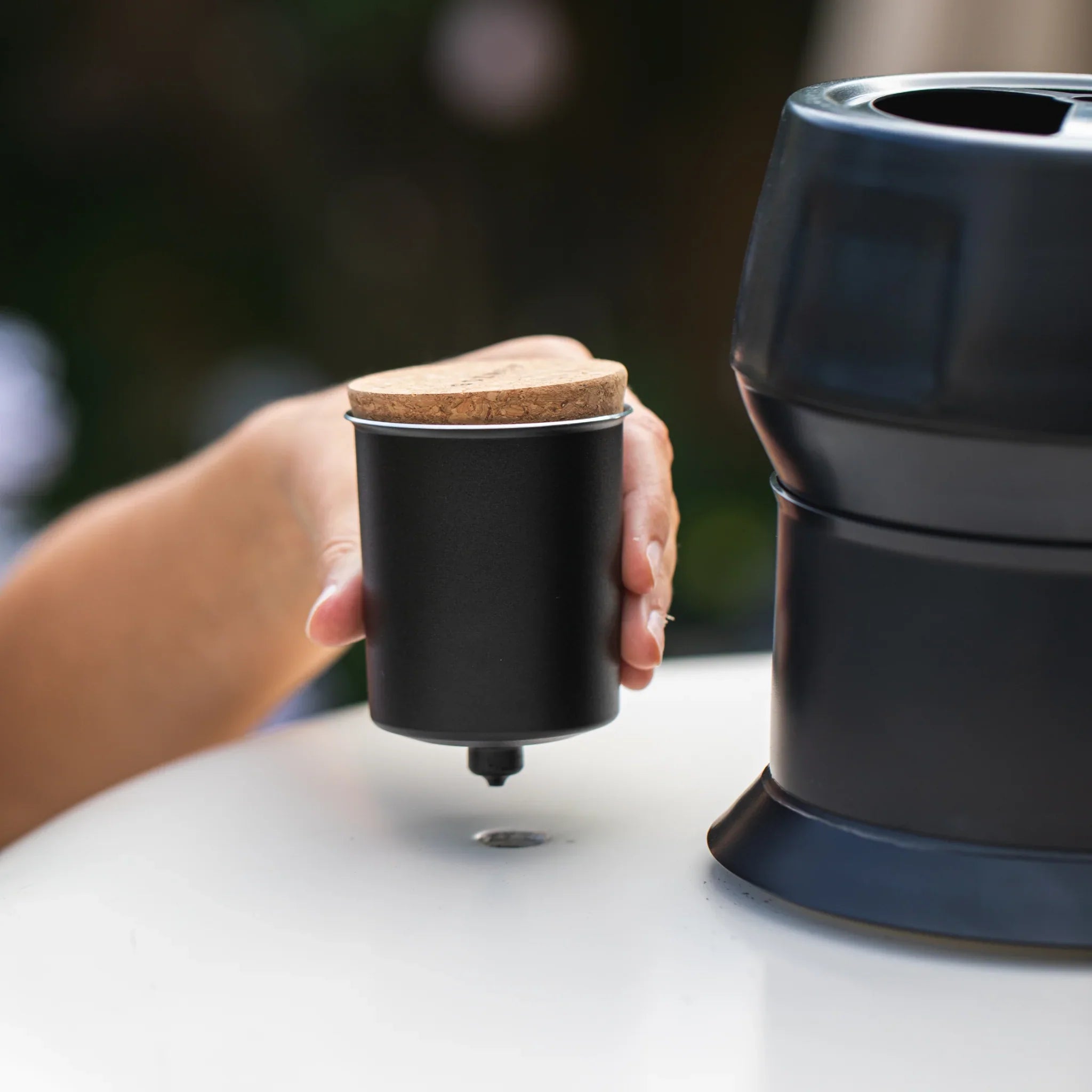 Person holding a black portable coffee maker with a cork lid on a blurred outdoor background